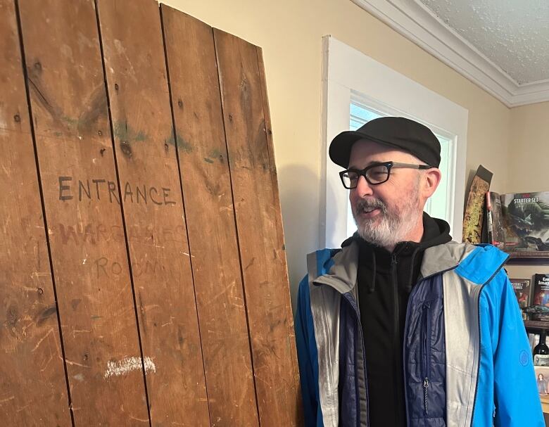 A man wearing a blue coat stands in a living room. He has a smile on his face as he stares at a large wooden door taken off it's hinges.