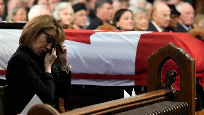 A woman wipes away tears, with a casket wrapped in a Canadian flag in the background.