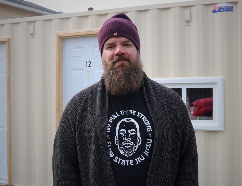 A bearded man in a burgundy toque stands in front of a shipping container. 