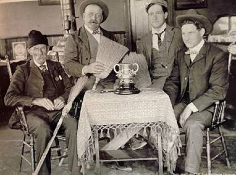 An old timey black and white photo shows four men sitting aound a trophy.