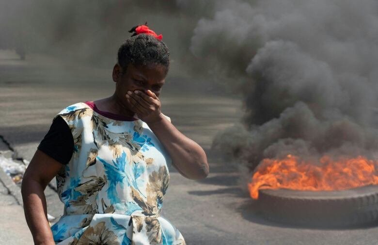 A woman with her hand over her mouth and nose walks down a street by a tire on fire. 