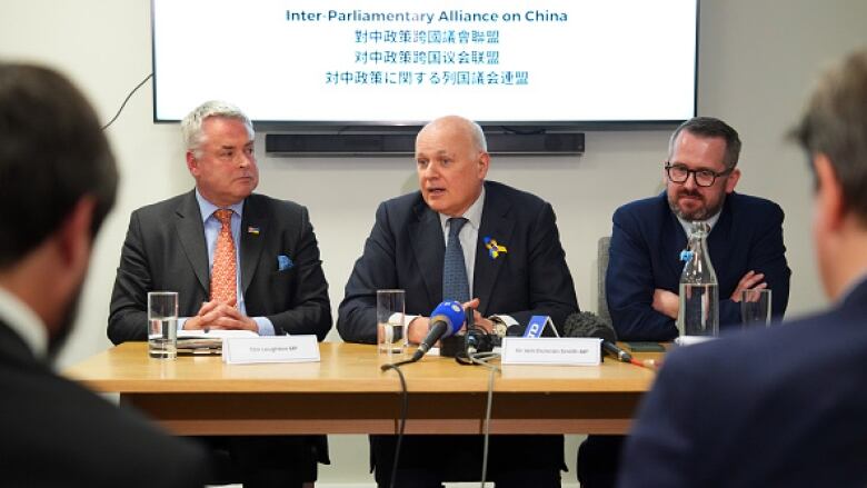 Three men in suits sit at a table with microphones placed in the centre.