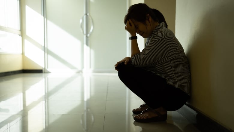 A woman rests her head on her hand as she crouches alone in a darkened room.