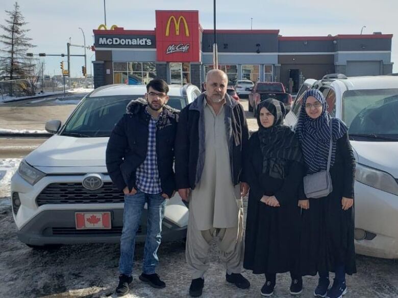 A group of four people stand next to a Toyota car with a Canadian flag licence plate. They are standing in front of a McDonald's restaurant.