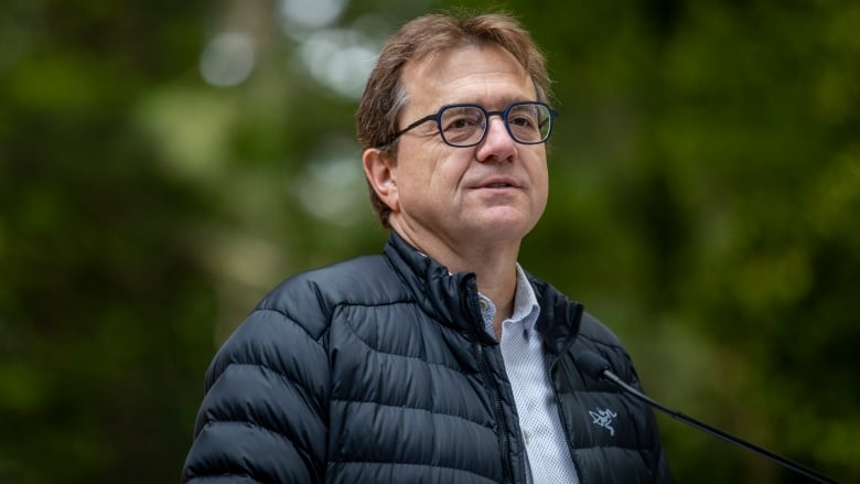 A white man wearing a black puffer jacket and blue glasses speaks to a mic while shrouded by trees.