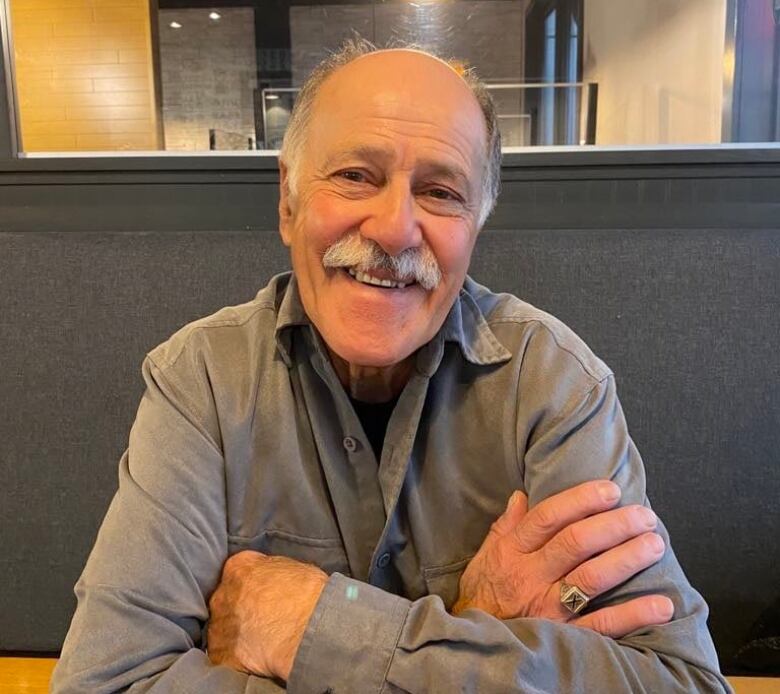 A man with greying hair and a grey moustache with a dark grey collared up shirt sits with arms folded and is smiling.