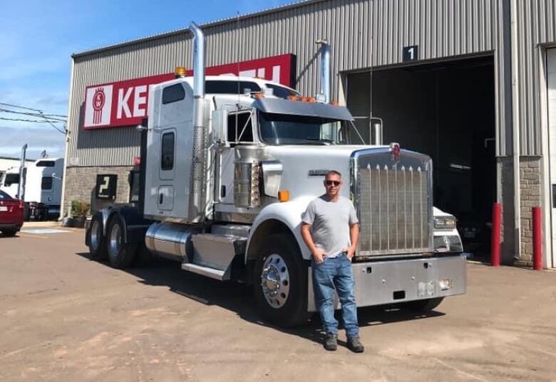 A man with a t-shirt and blue jeans stands in front of the cab of a transport truck which is parked in front of a garage.