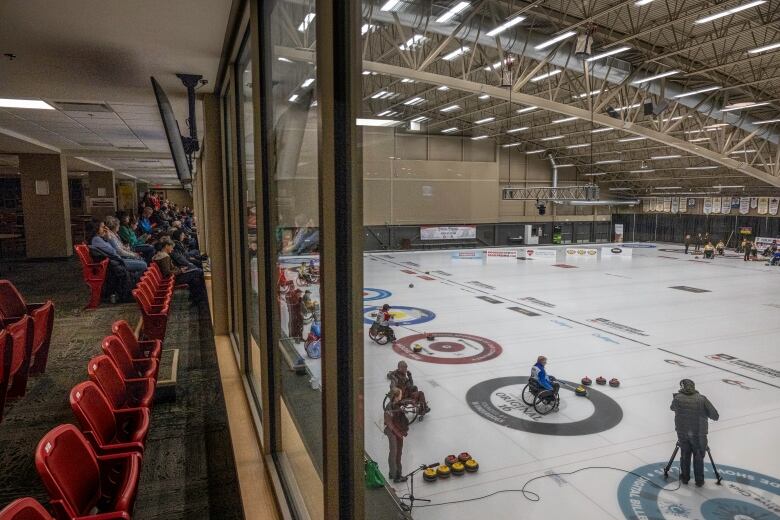 A group of people sit in viewers' seats watching teams do wheelchair curling.