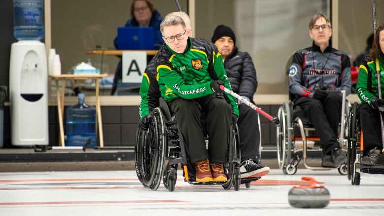 A man in a wheelchair wearing a green uniform sends a curling rock down the ice.