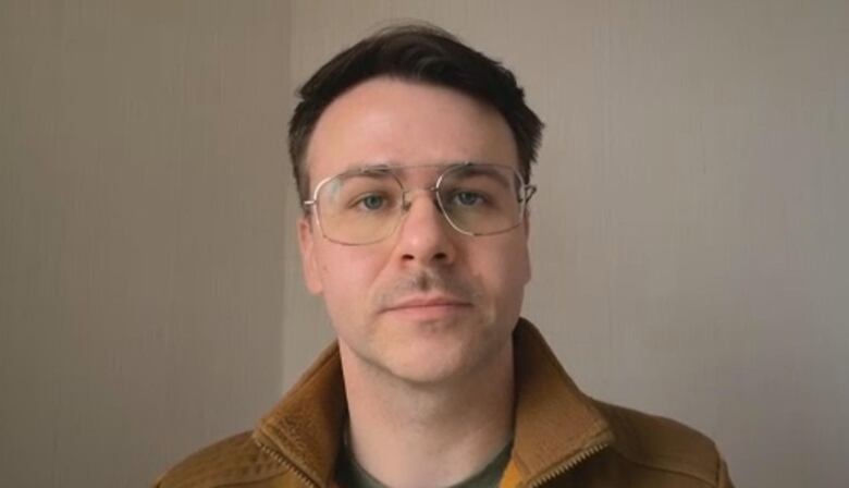 A photo of Kent Peterson against a beige-grey wall wearing a mustard yellow jacket, green t-shirt and thin frame eye glasses. 