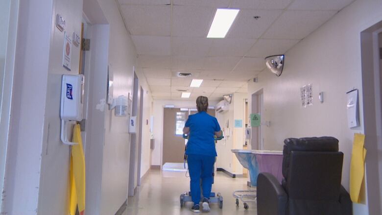 A nurse wearing blue scrubs and sneakers pushes medical equipment down a hallway.