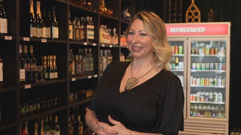 A woman with blonde hair and wearing a black dress is smiling. Behind her are shelves and a glass refrigerator lined with non-alcoholic drinks