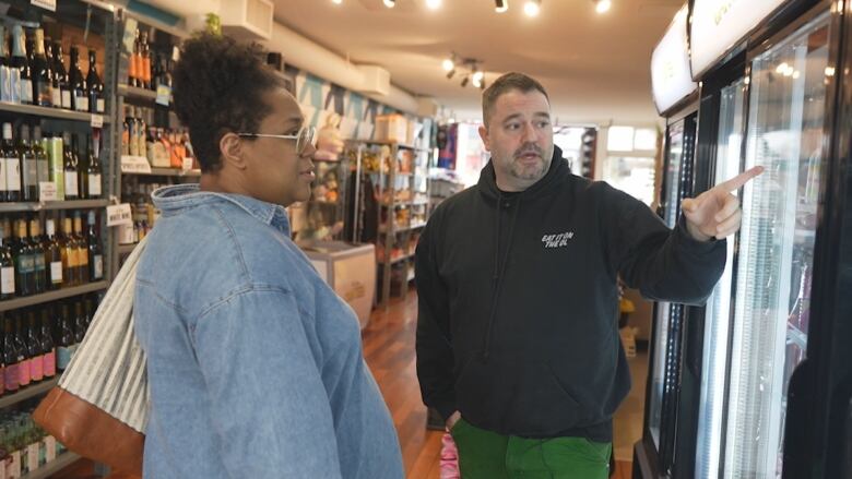 A man in a blue hoodie is pointing into a glass refrigerator and speaking to a woman in a blue shirt and holding a striped brown bag. 