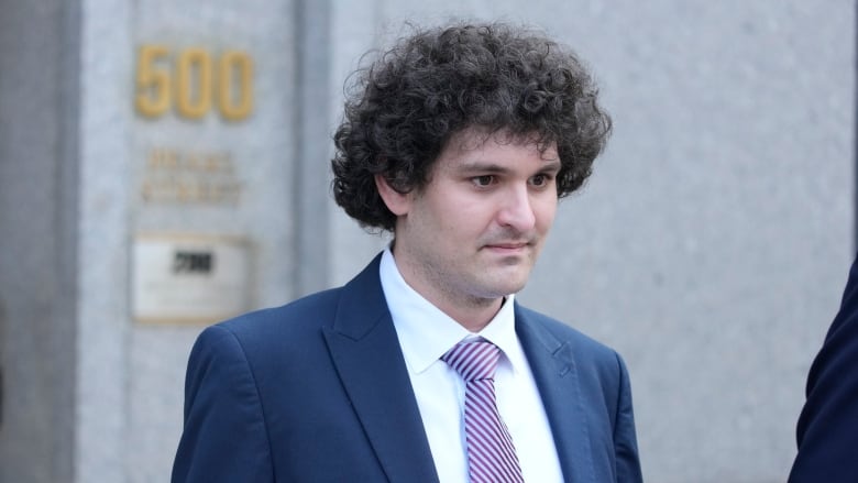 A cleanshaven person with thick, curly hair wearing a suit and tie is shown standing outside.