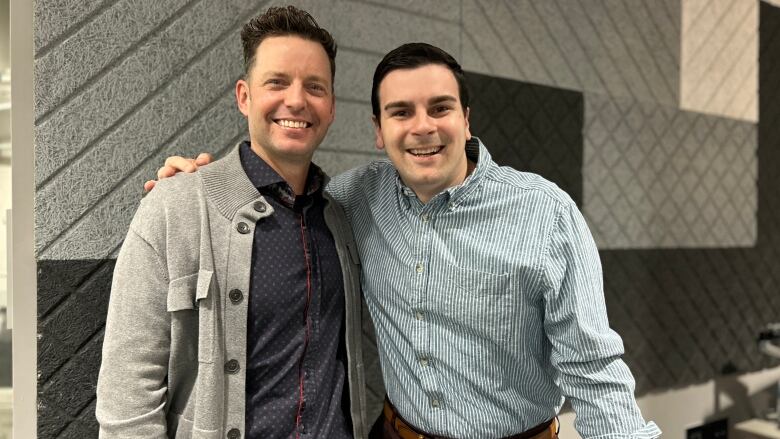 Two men standing in front of a grey wall.