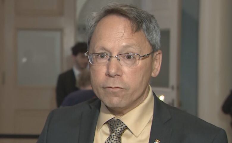 A man wearing a suit, tie and glasses stands in a hallways.