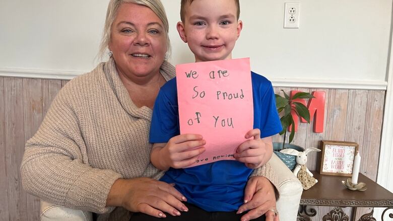 A woman with blonde hair sits on a couch with a six year old child, who holds a hand-drawn card reading 'We are so proud of you.'