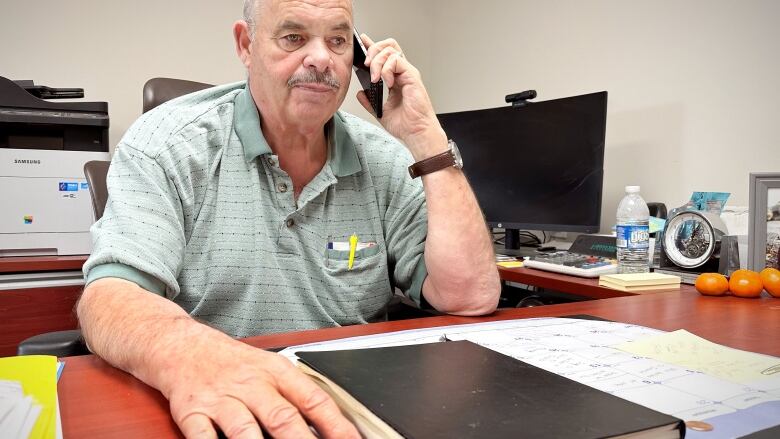 A man in a celadon polo shirt  speaks on the phone while sitting at a desk.