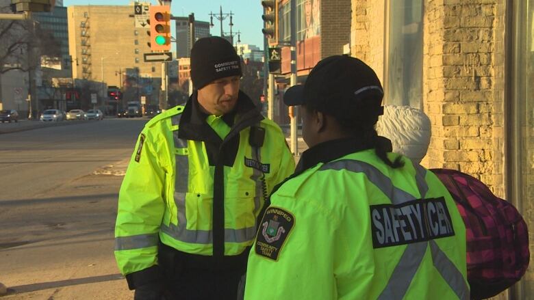 A man wearing a yellow and black jacket is speaking to someone standing behind a woman wearing the same jacket, standing with her back to the camera.