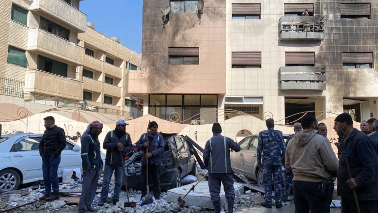 Several men with broom are shown in a city square near pieces of concrete debris and damaged vehicles and buildings.