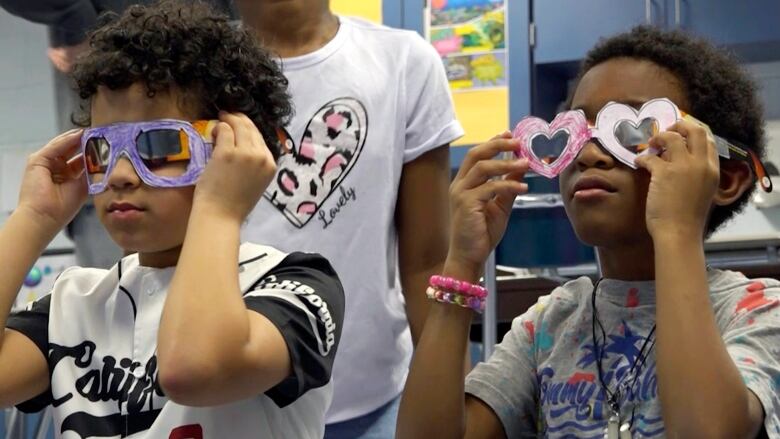 Two young children hold up glasses they made from paper. 