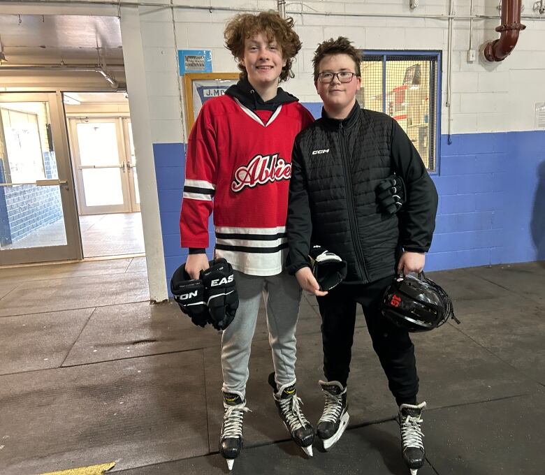 Two young boys stand together with skates on inside an ice arena