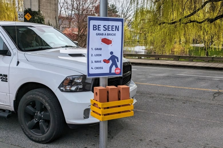 A sign reads 'Be Seen, Grab a Brick' with bricks underneath it in a holder. A car crosses a crosswalk in the background.