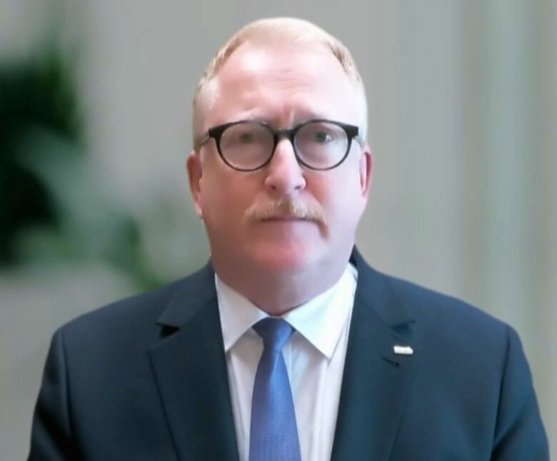 A man wearing glasses and a black suit sitting in an office. 