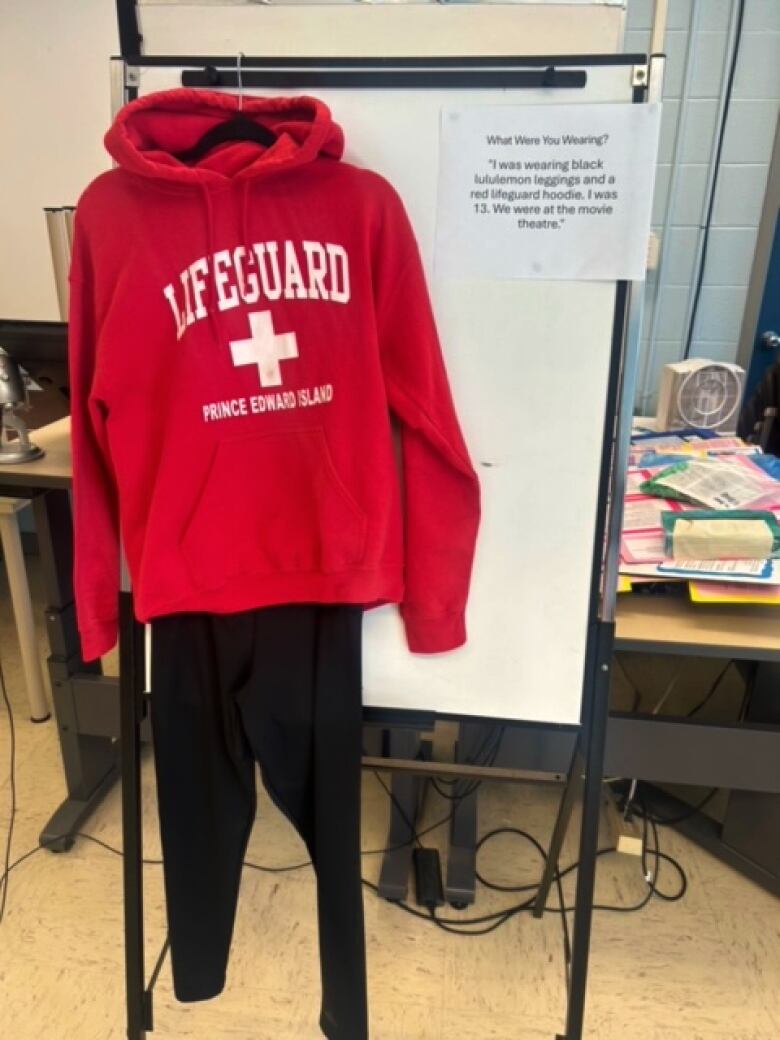 A red hoodie with the word lifeguard is hung up with a pair of black leggings. A piece of paper beside it reads 