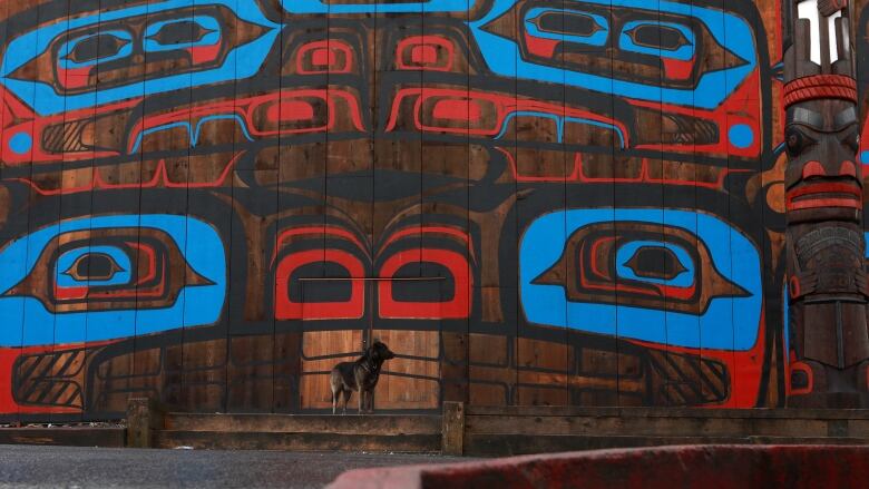 A dog sits in front of a large building with Indigenous art.