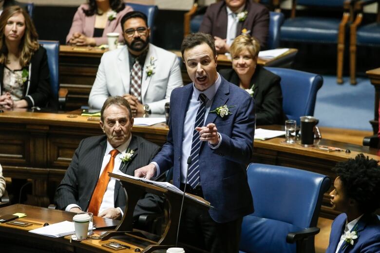 A man who is standing points as he speaks toward the camera in a large chamber in a legislative building.