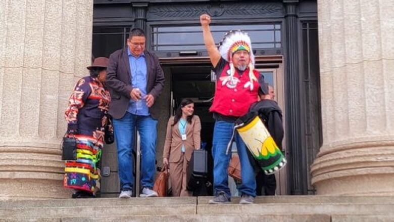 A man wearing a headdress raises his fist in the air on the steps of a building.