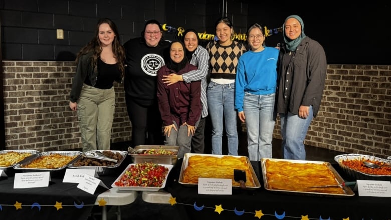 Seven women, including three wearing traditional headscarves, pose behind a series of tables that are loaded with food.