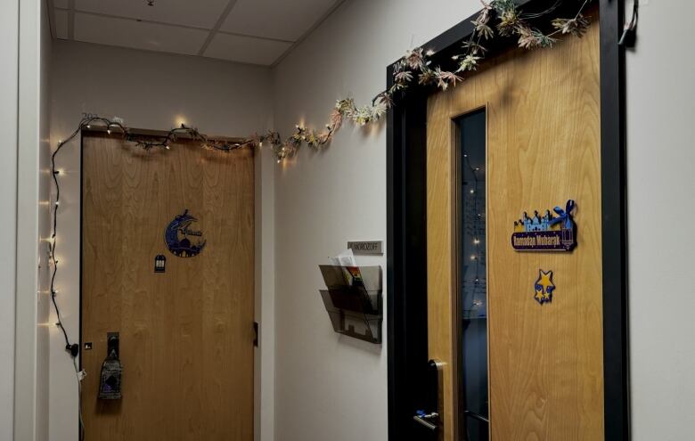 An office hallway with two wooden doors decorated with lights, garland and signs that read Ramadan Mubarak and Eid Mubarak.