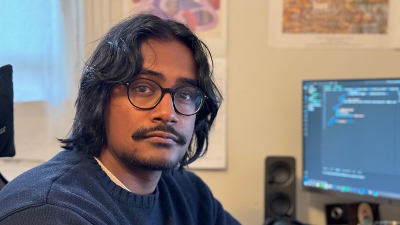 A man wearing glasses and a blue sweater sits at a desk with a computer monitor seen in the background.