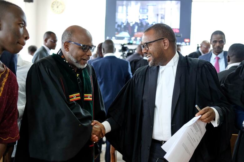 Two bald men in black robes smile and shake hands.