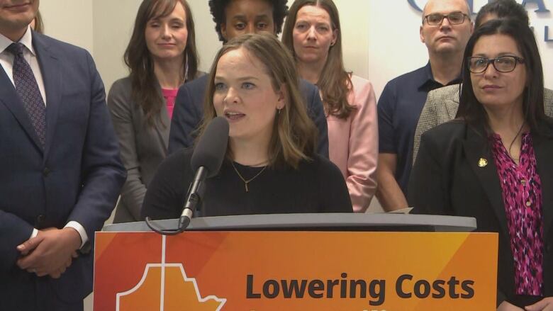 A woman with shoulder-length brown hair in a black shirt speaks into a microphone at a lectern during an government health-care announcement.