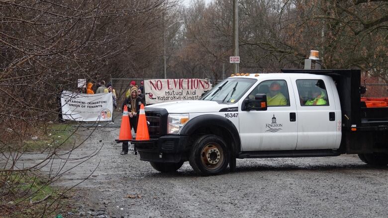 A white truck being driven by a man with an orange hard hat and with the City of Kingston logo on the door turns on a gravel road. Behind the truck is a group of people blocking a gate with a banner that reads 