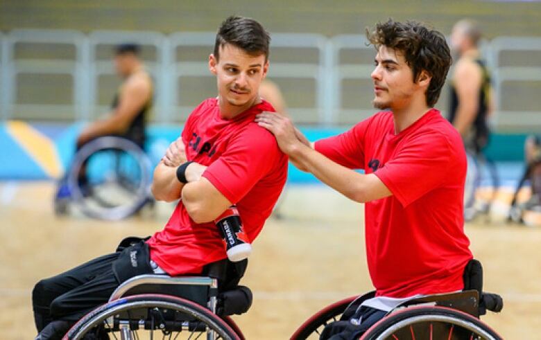 Two men wearing red shirts sit in wheelchairs on a basketball court.