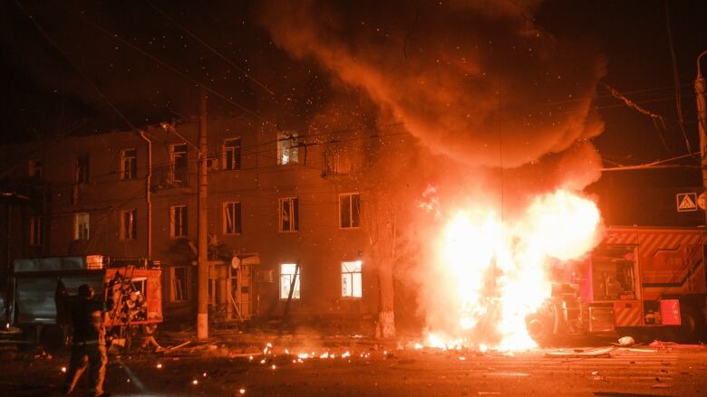 A large ball of fire is shown bursting forth from a building in a nighttime photograph. A firefighter is seen across the street.