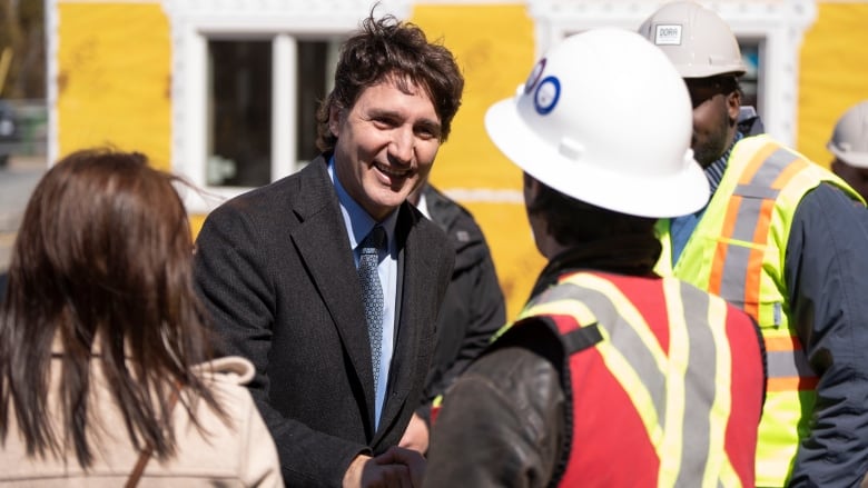 A man in dark hair and a suit smiles and shakes hands with people in hard hats and construction vests