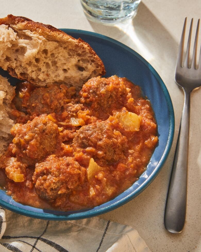 Closeup on a shallow blue bowl with Sweet-and-Sour Meatballs and a piece of bread in it. A fork sits next to the bowl. 