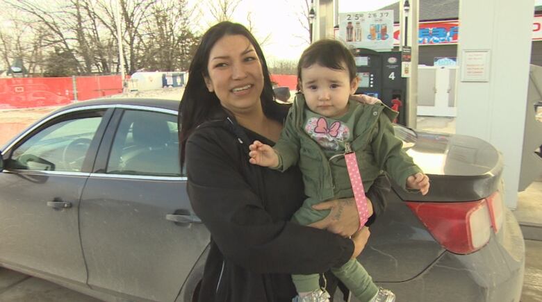 A woman with long, dark hair holds a small child in front of her car at a gas station.