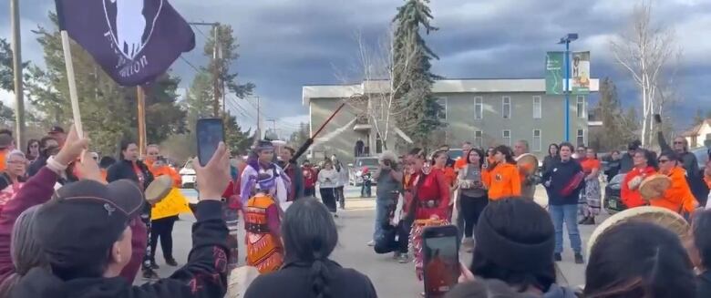 A crowd of people, some holding cell phones, flags, or drums, form a circle around Indigenous dancers wearing regalia, during a protest march to city hall.  