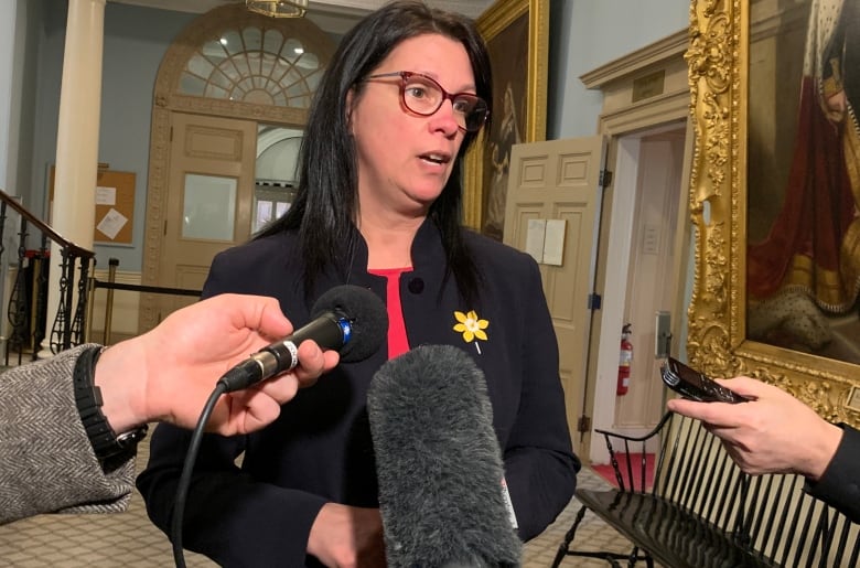 A woman with glasses stands in front of microphones.