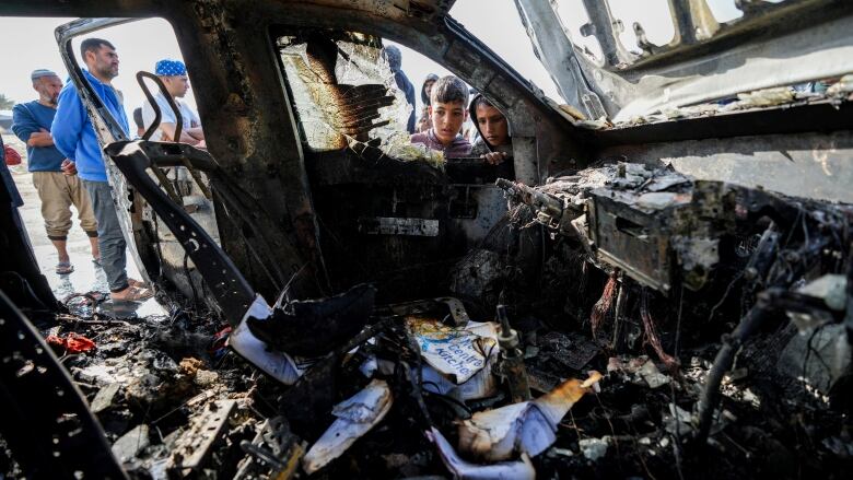 A child looks through a shattered window into the charred remains of a van with a visible World Central Kitchen logo. 