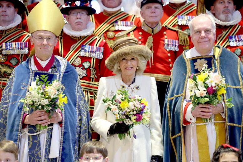 A person holding a bouquet of flowers stands betwee two people in religious vestments also holding bouquets of flowers and in front of retired service members in ceremonial dress.