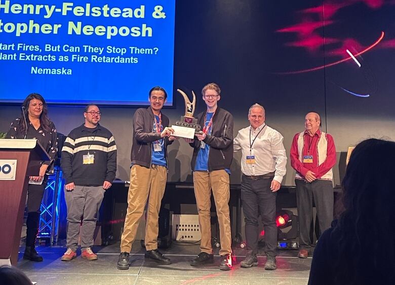 Two teenaged boys stand on a stage holding a trophy, with other people standing beside them.