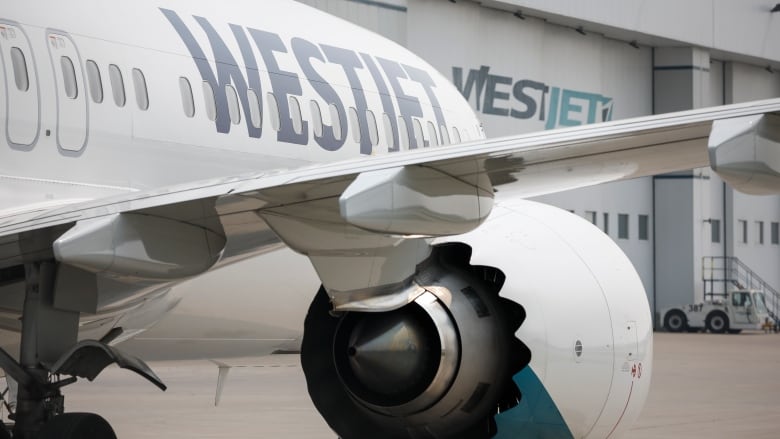 The wing and front portion of a passenger plane with the name WesJet is seen outside of a hangar with the WestJet name as well. 