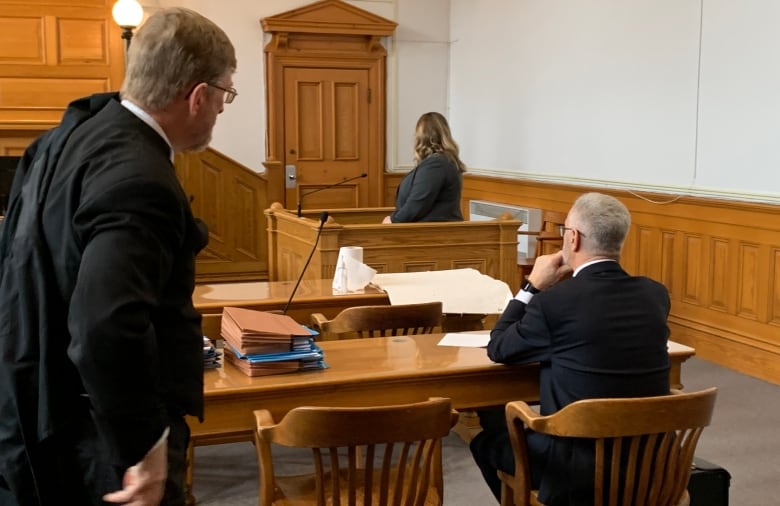 A woman in a grey suit stands in a wooden box in front of a microphone. A greyhaired man is sitting facing her, wearing a black suit. 